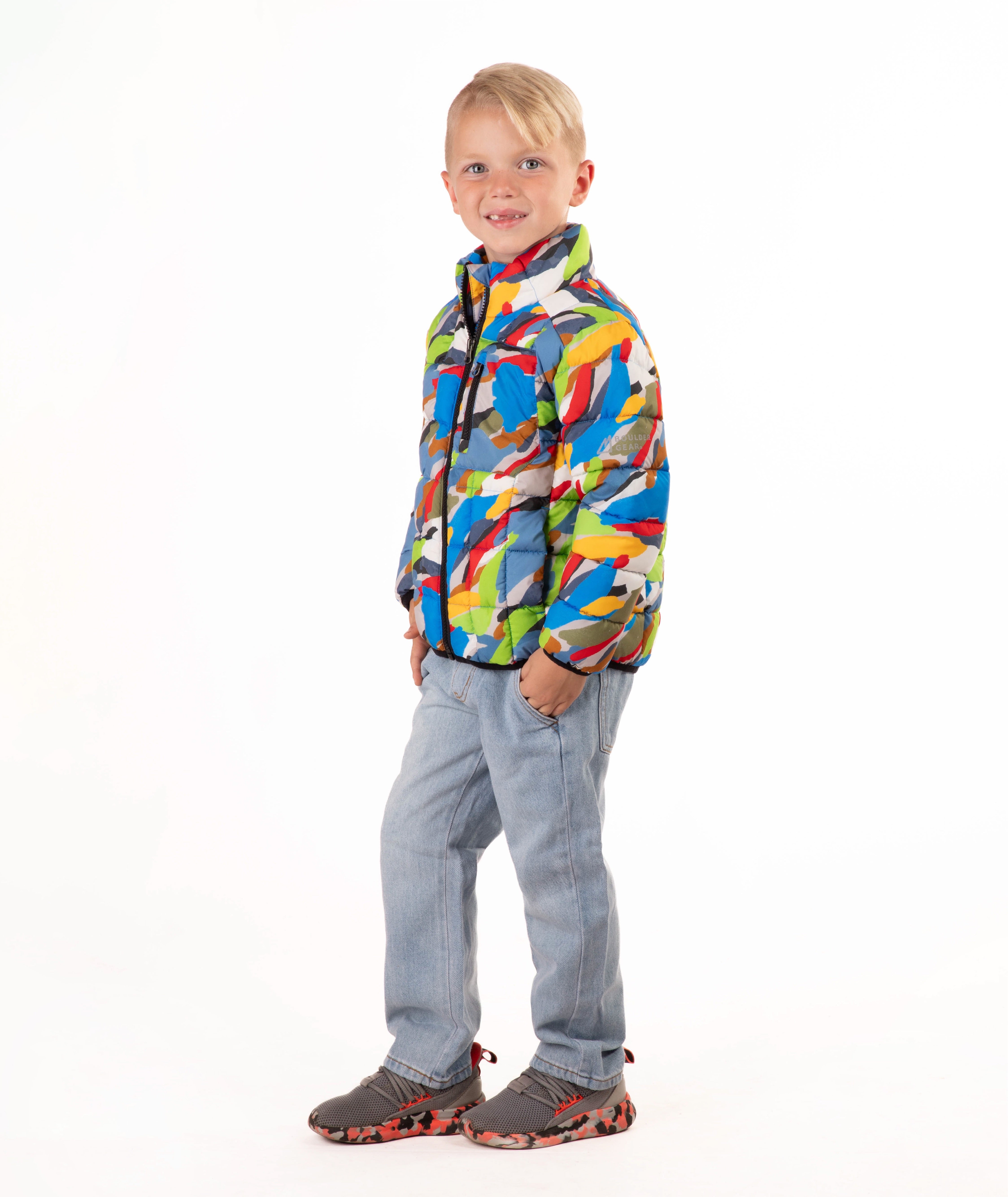 A young boy stands against a white studio background wearing a colorful puffer jacket with a multi color camo print, blue jeans, and brown shoes.