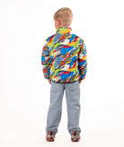 A young boy stands against a white studio background wearing a colorful puffer jacket with a multi color camo print, blue jeans, and brown shoes.