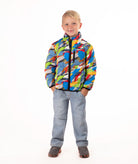 A young boy stands against a white studio background wearing a colorful puffer jacket with a multi color camo print, blue jeans, and brown shoes.