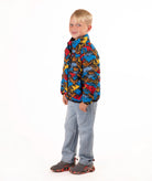 A young boy stands against a white studio background wearing a colorful puffer jacket with a multi color mountain print, blue jeans, and brown shoes.