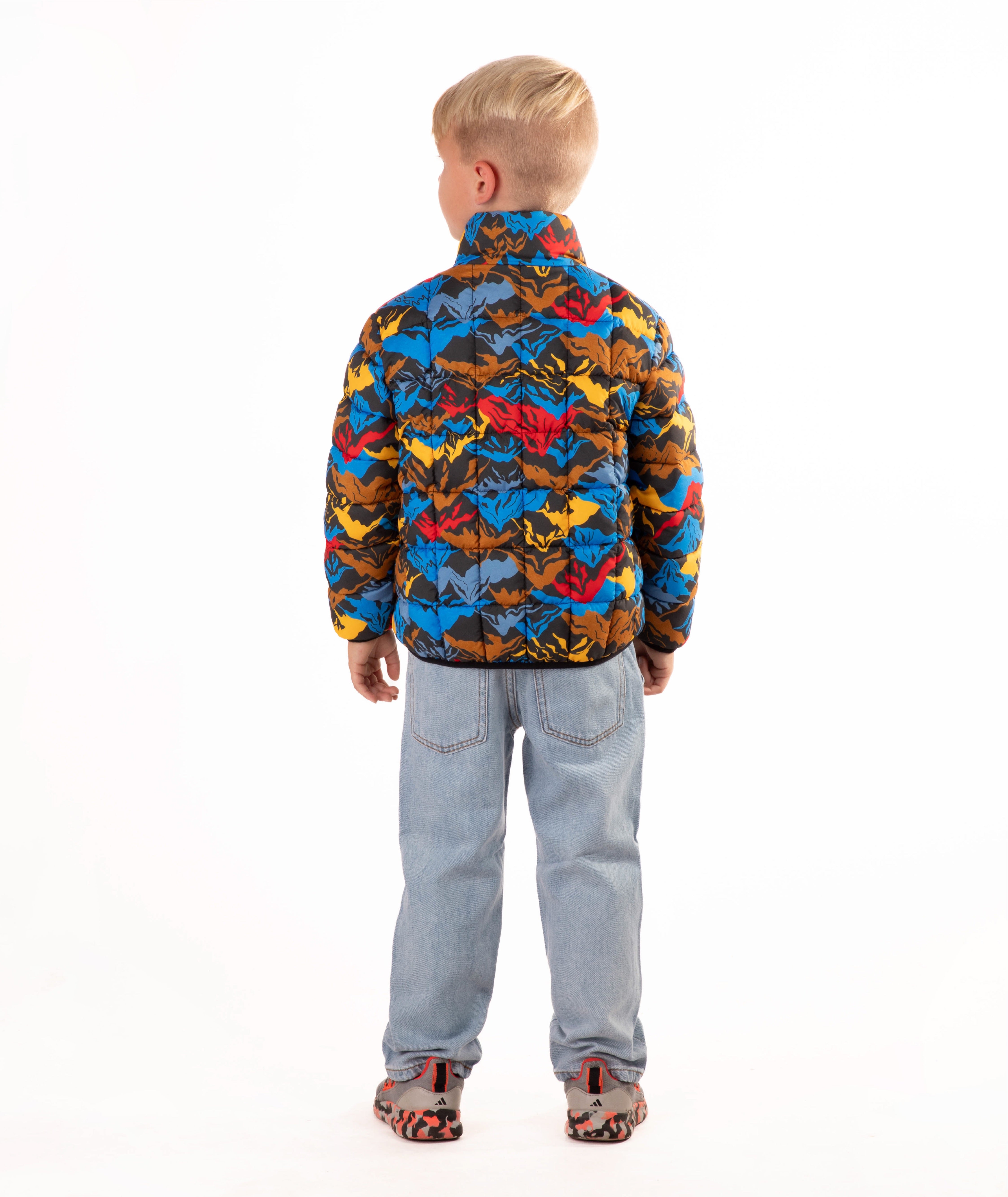 A young boy stands against a white studio background wearing a colorful puffer jacket with a multi color mountain print, blue jeans, and brown shoes.