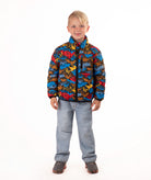 A young boy stands against a white studio background wearing a colorful puffer jacket with a multi color mountain print, blue jeans, and brown shoes.