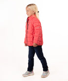 A young girl with blonde hair stands against a white studio background wearing a coral pink quilted puffer jacket, dark blue jeans, and gray sneakers.