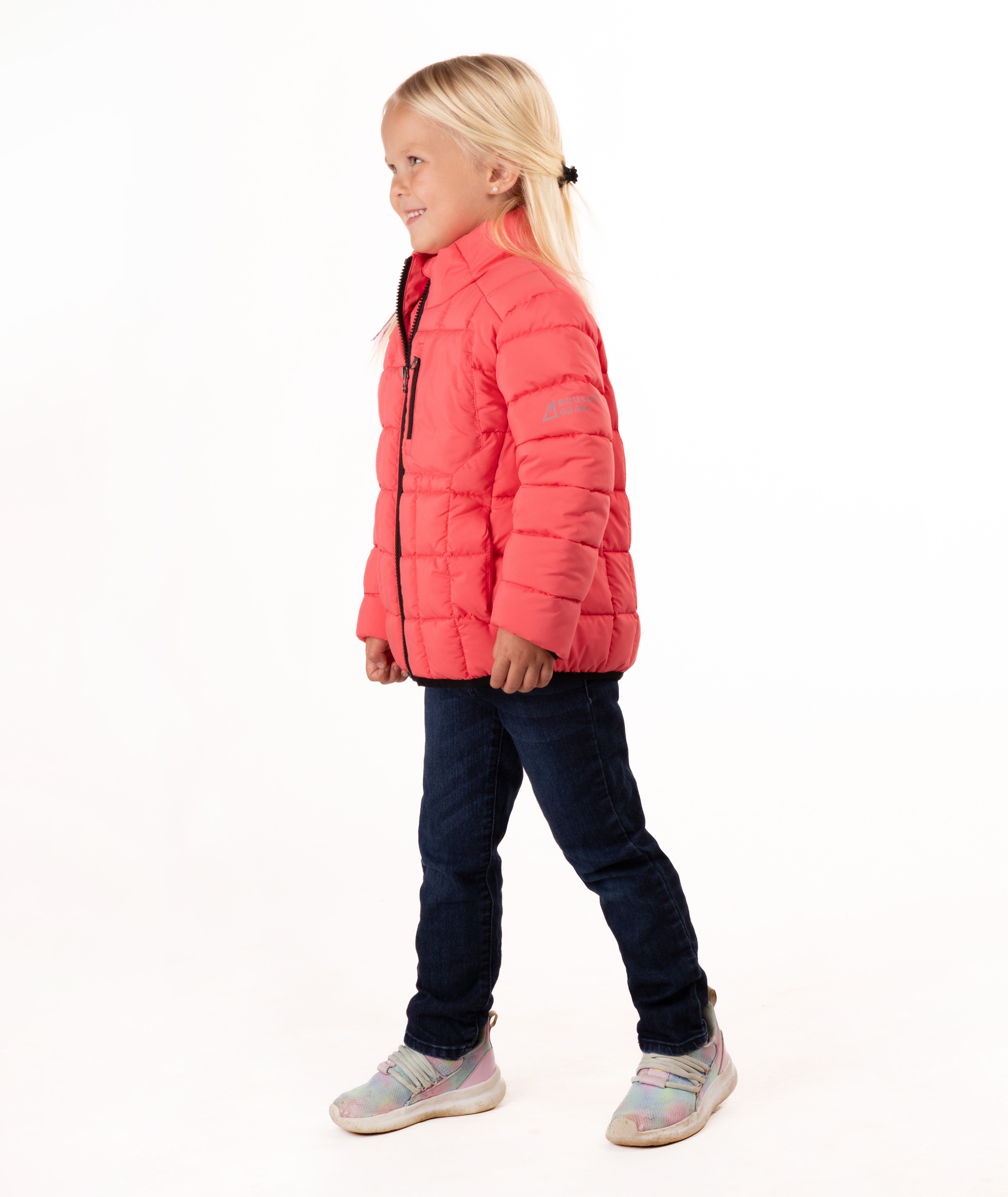 A young girl with blonde hair stands against a white studio background wearing a coral pink quilted puffer jacket, dark blue jeans, and gray sneakers.