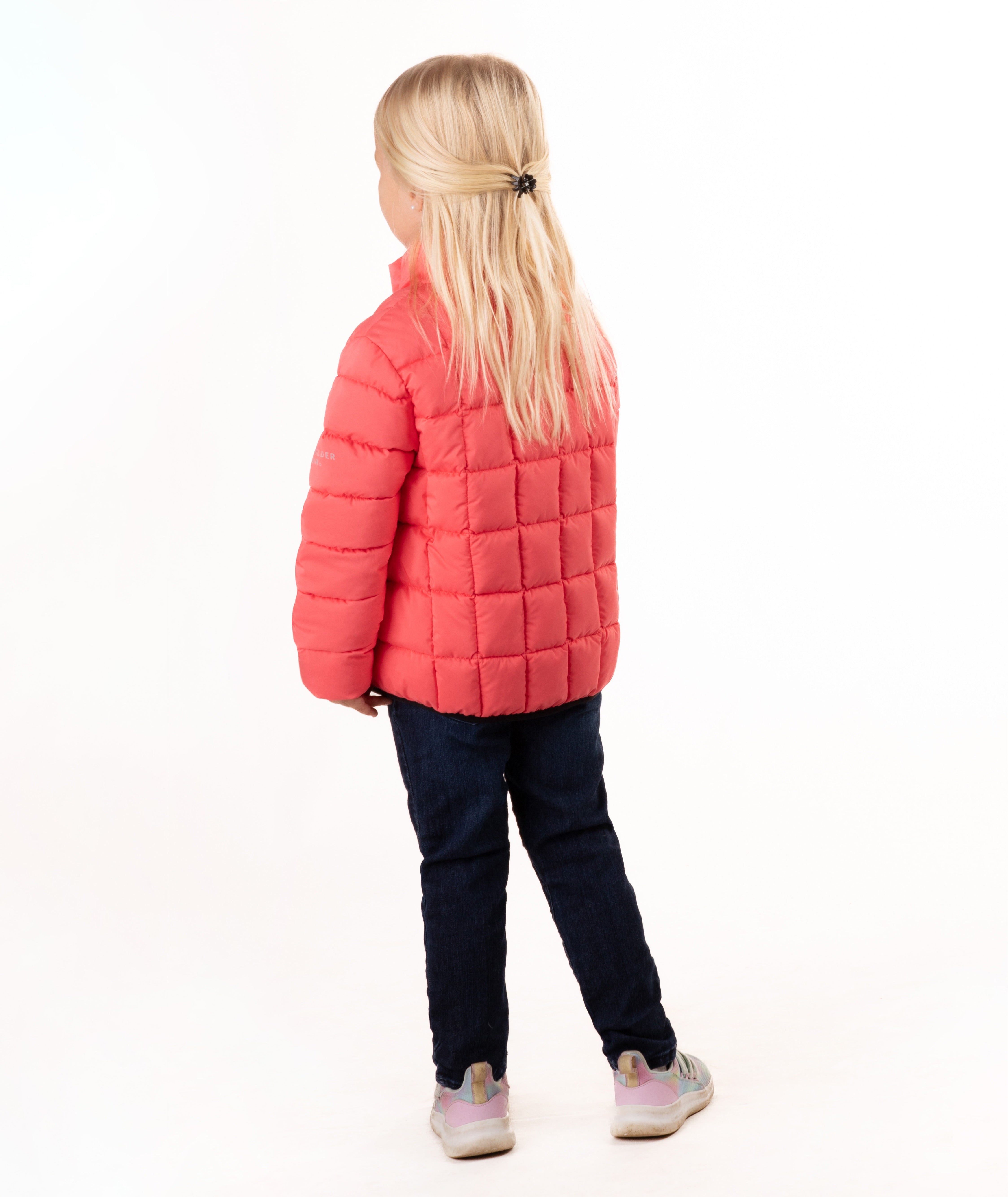 A young girl with blonde hair stands against a white studio background wearing a coral pink quilted puffer jacket, dark blue jeans, and gray sneakers.