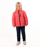 A young girl with blonde hair stands against a white studio background wearing a coral pink quilted puffer jacket, dark blue jeans, and gray sneakers.