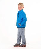 A young boy wearing a bright blue quilted puffer jacket, light blue jeans, and brown sneakers, standing against a solid white background.