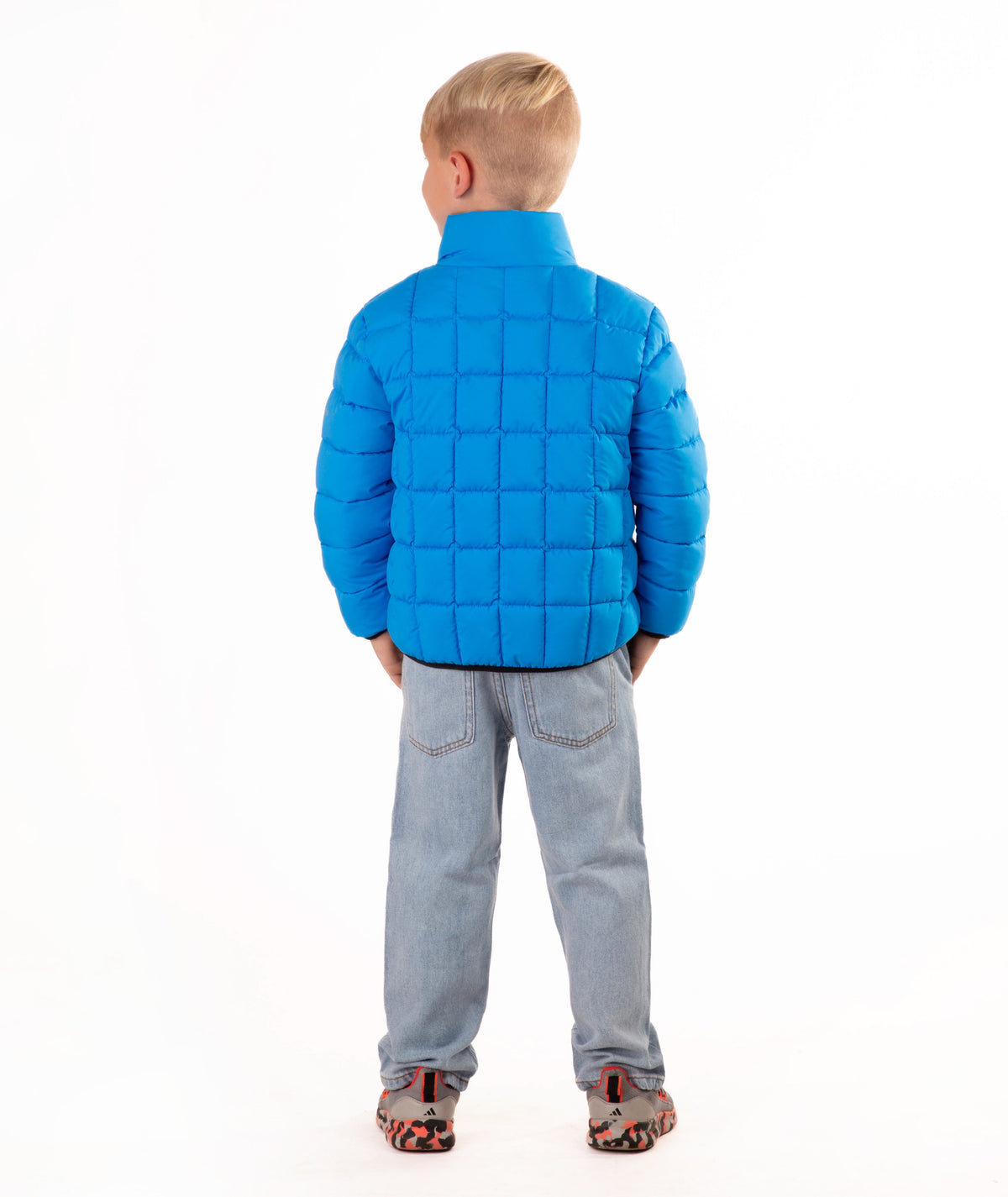 A young boy wearing a bright blue quilted puffer jacket, light blue jeans, and brown sneakers, standing against a solid white background.