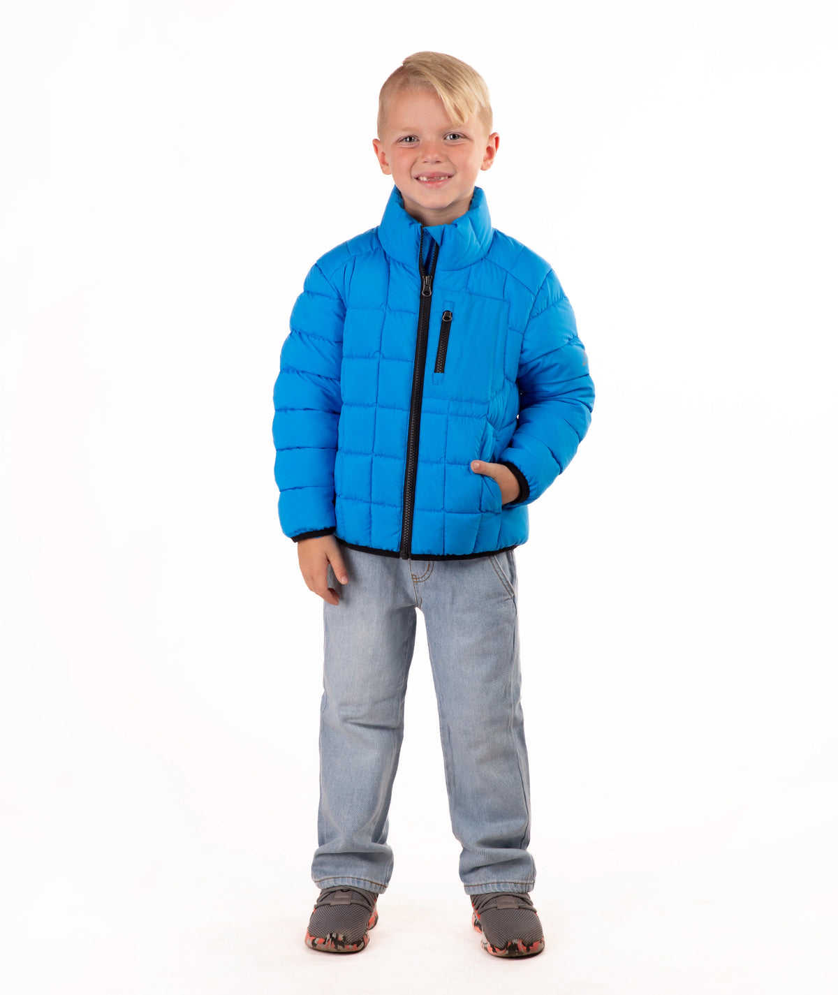 A young boy wearing a bright blue quilted puffer jacket, light blue jeans, and brown sneakers, standing against a solid white background.