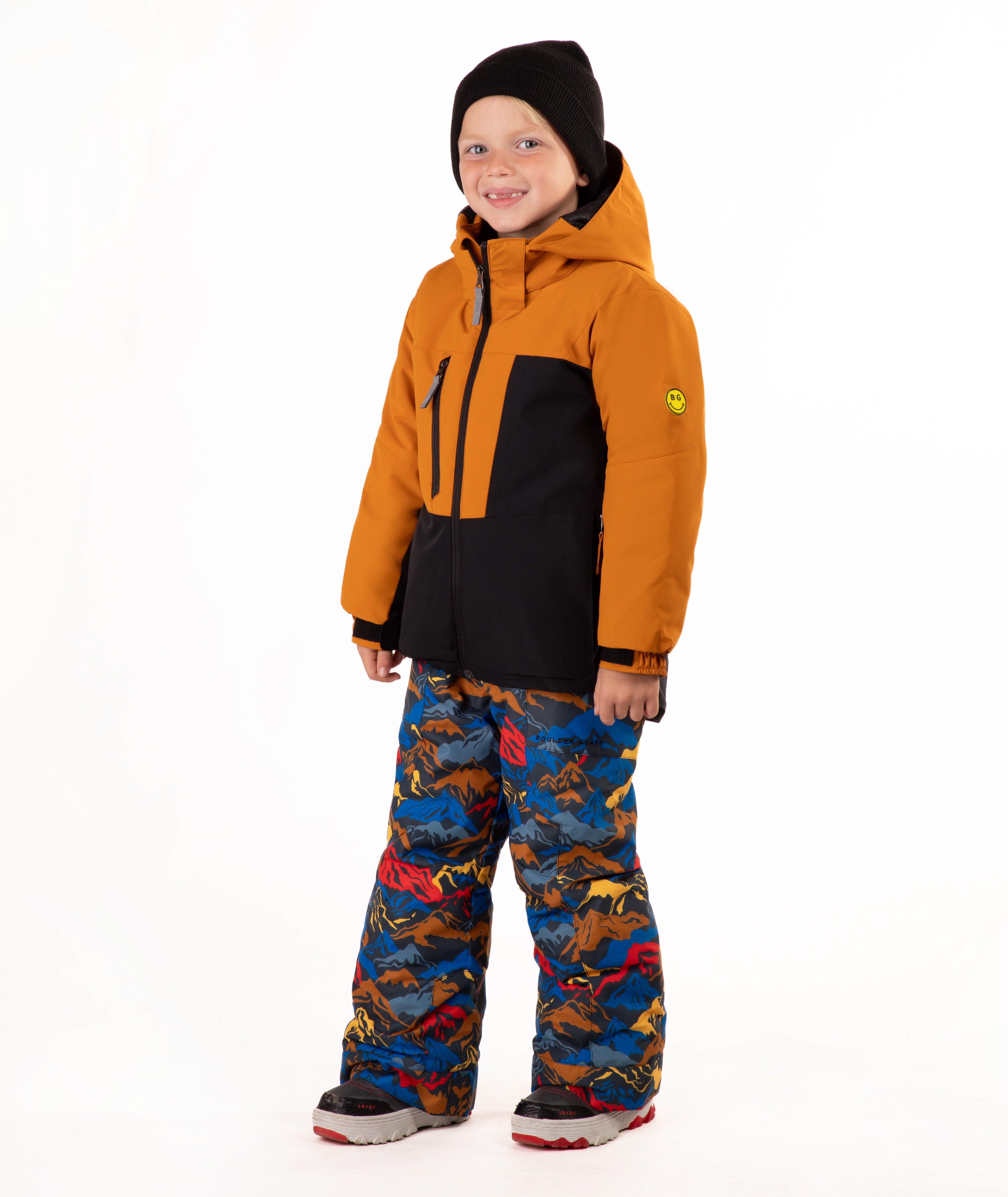 A child wearing a kids' ski jacket and snow pants set against a plain white background. The jacket is a colorblock design in brown and black, paired with blue snow pants featuring a multicolor mountain and ski pattern.