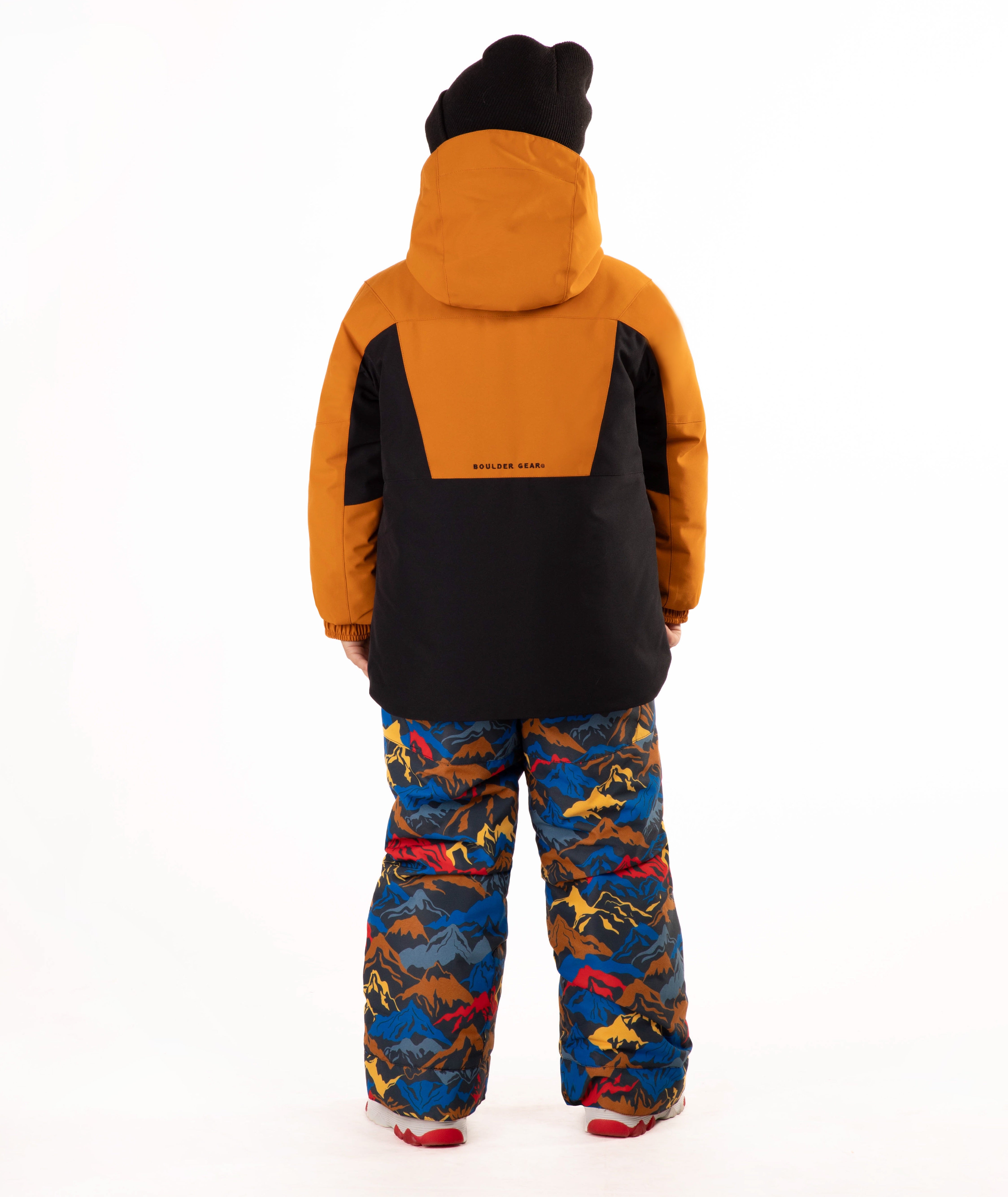 A child wearing a kids' ski jacket and snow pants set against a plain white background. The jacket is a colorblock design in brown and black, paired with blue snow pants featuring a multicolor mountain and ski pattern.