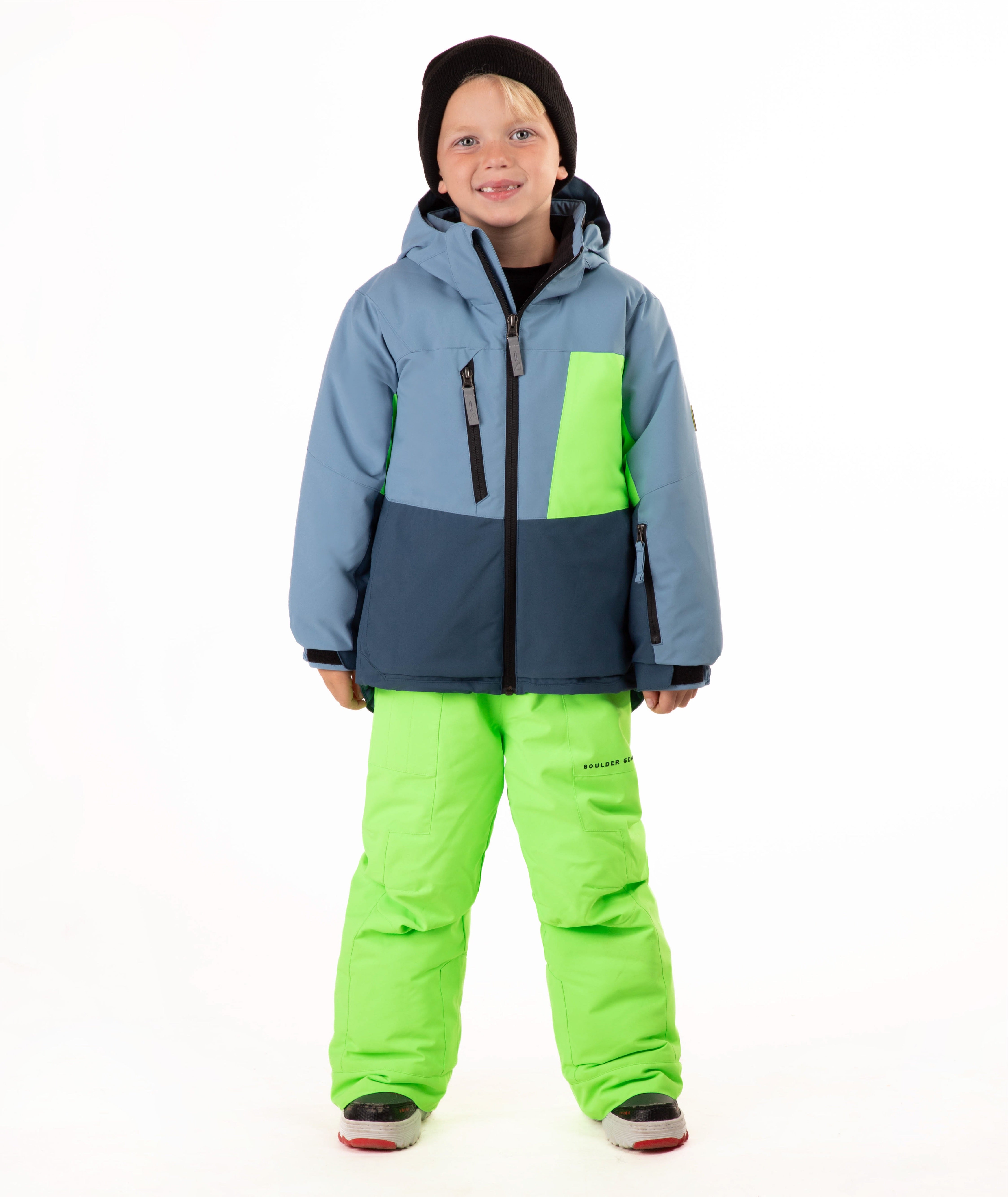 A child wearing a color-blocked ski jacket in slate blue, navy, and neon green, paired with neon green ski pants, a black beanie, standing against a white studio background.