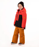 A young child wearing a red and black hooded snow jacket, rust-orange snow pants, a black beanie, standing against a plain white studio background.