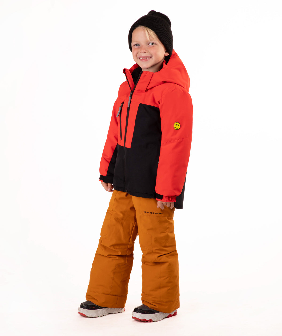 A young child wearing a red and black hooded snow jacket, rust-orange snow pants, a black beanie, standing against a plain white studio background.