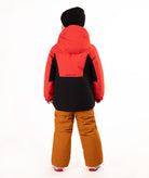 A young child wearing a red and black hooded snow jacket, rust-orange snow pants, a black beanie, standing against a plain white studio background.