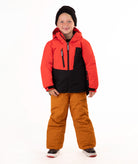 A young child wearing a red and black hooded snow jacket, rust-orange snow pants, a black beanie, standing against a plain white studio background.