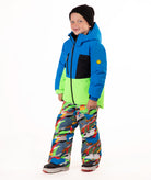 A child wearing a bright blue and green ski jacket with a black chest panel, colorful camouflage-patterned snow pants, and a black beanie, standing against a plain white background.