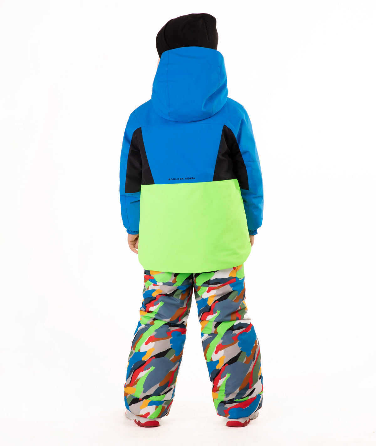A child wearing a bright blue and green ski jacket with a black chest panel, colorful camouflage-patterned snow pants, and a black beanie, standing against a plain white background.