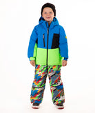 A child wearing a bright blue and green ski jacket with a black chest panel, colorful camouflage-patterned snow pants, and a black beanie, standing against a plain white background.