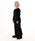 A child wearing black snow bib overalls over a black long-sleeve shirt and black beanie, standing against a plain white studio background.