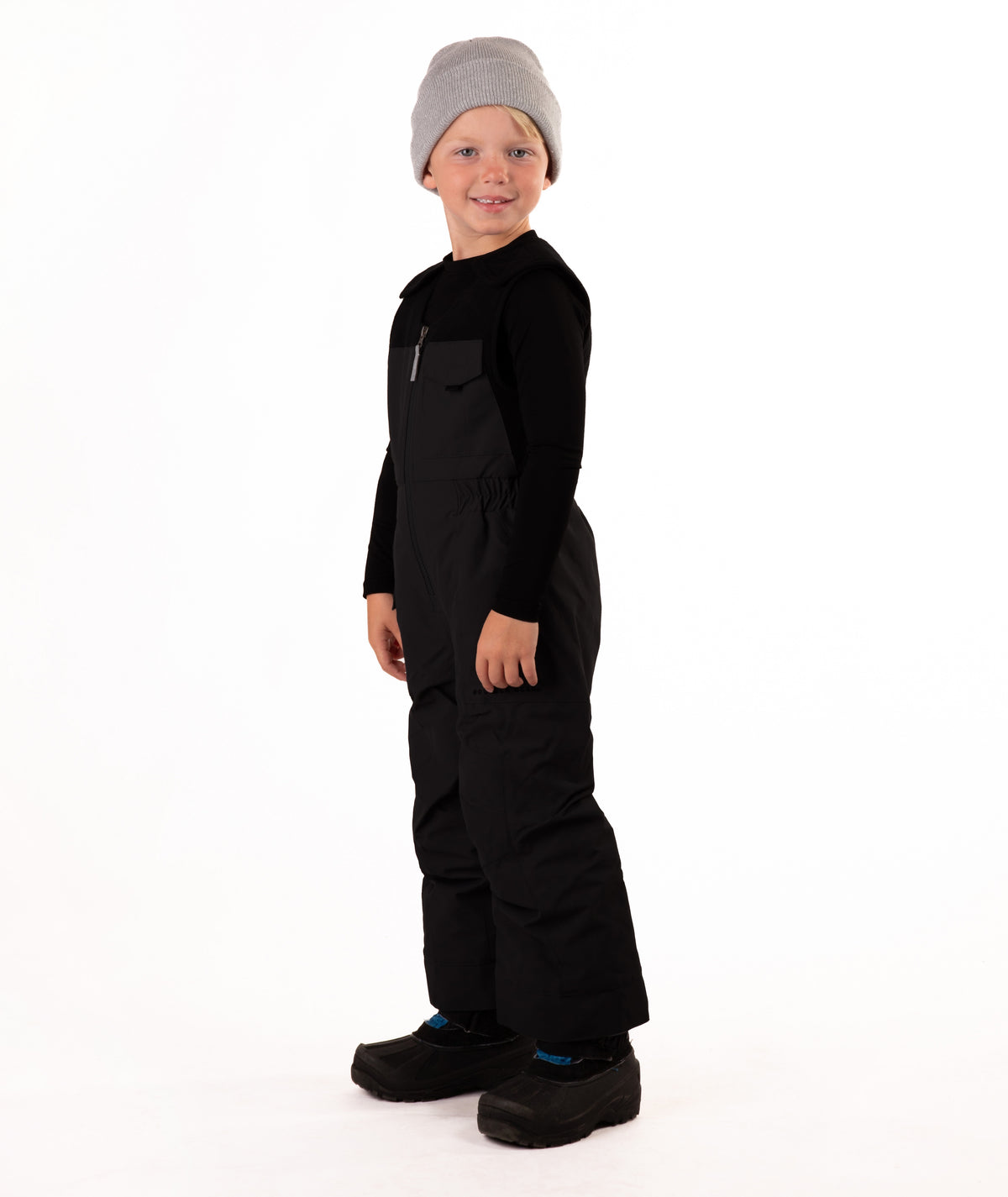 A child wearing black snow bib overalls over a black long-sleeve shirt and black beanie, standing against a plain white studio background.