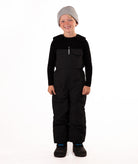A child wearing black snow bib overalls over a black long-sleeve shirt and black beanie, standing against a plain white studio background.