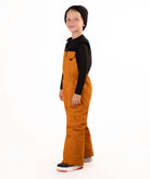 A child wearing a black knit beanie, a black long-sleeve top, and rust colored snow bib overalls, standing against a plain white background.