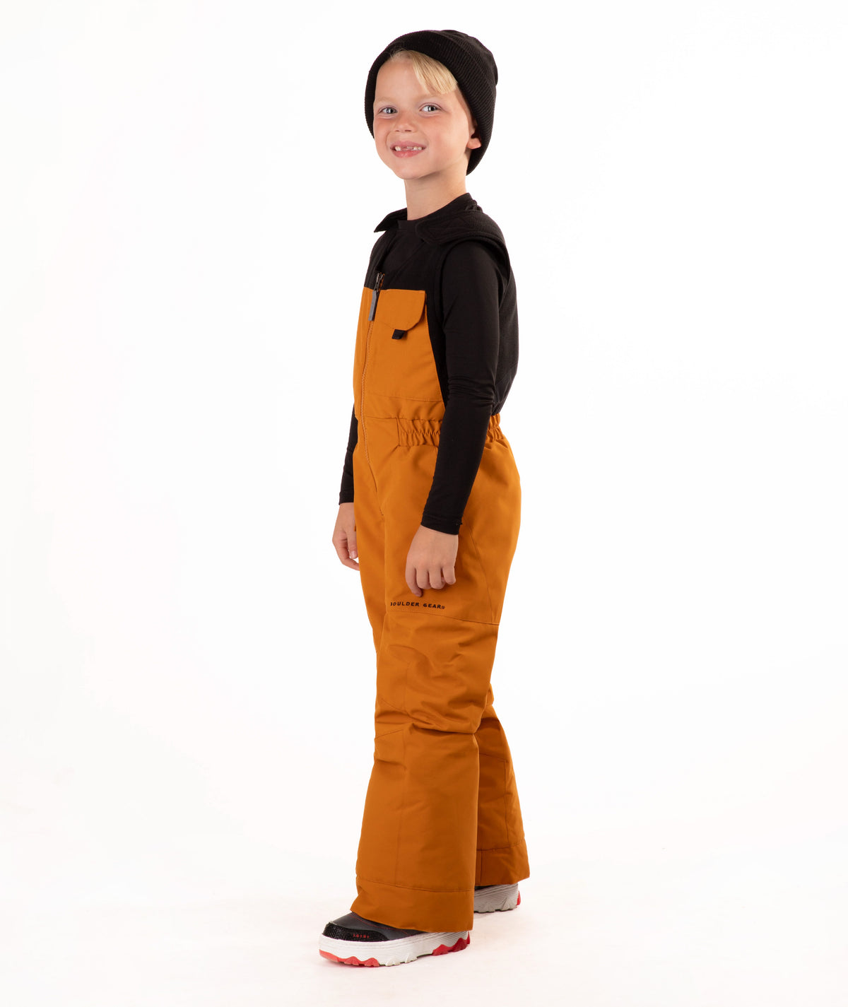 A child wearing a black knit beanie, a black long-sleeve top, and rust colored snow bib overalls, standing against a plain white background.