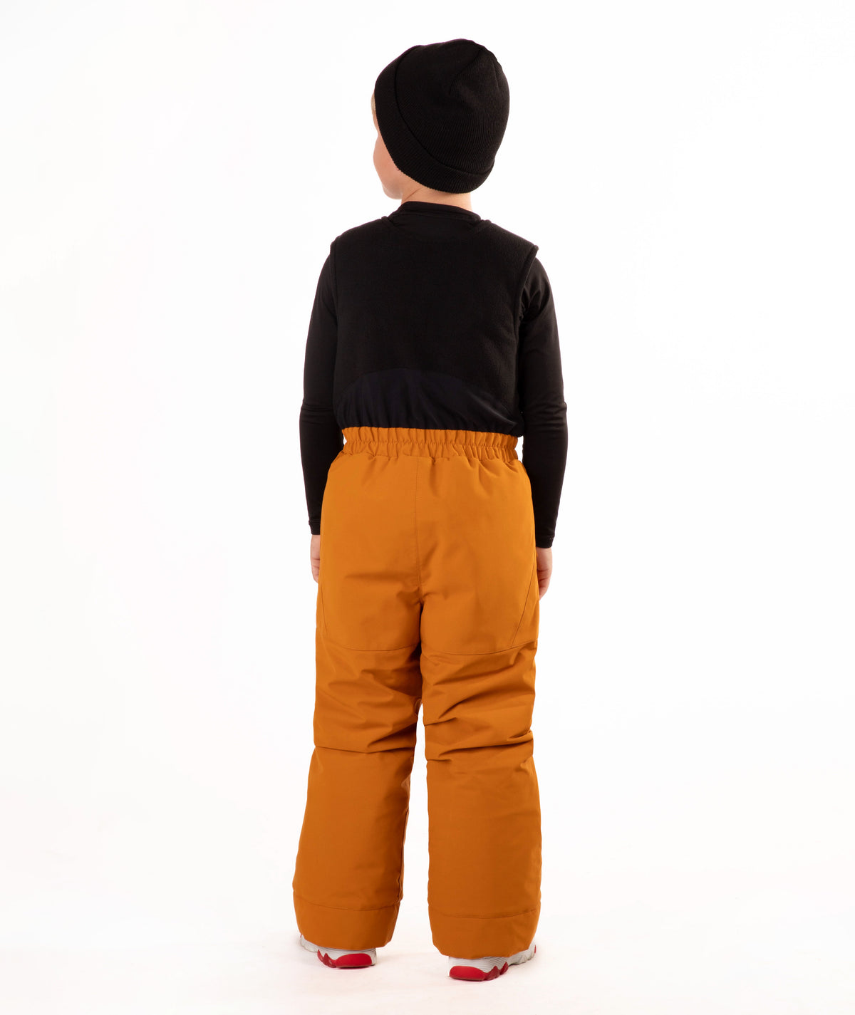 A child wearing a black knit beanie, a black long-sleeve top, and rust colored snow bib overalls, standing against a plain white background.