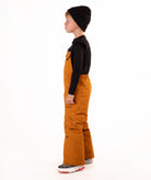 A child wearing a black knit beanie, a black long-sleeve top, and rust colored snow bib overalls, standing against a plain white background.