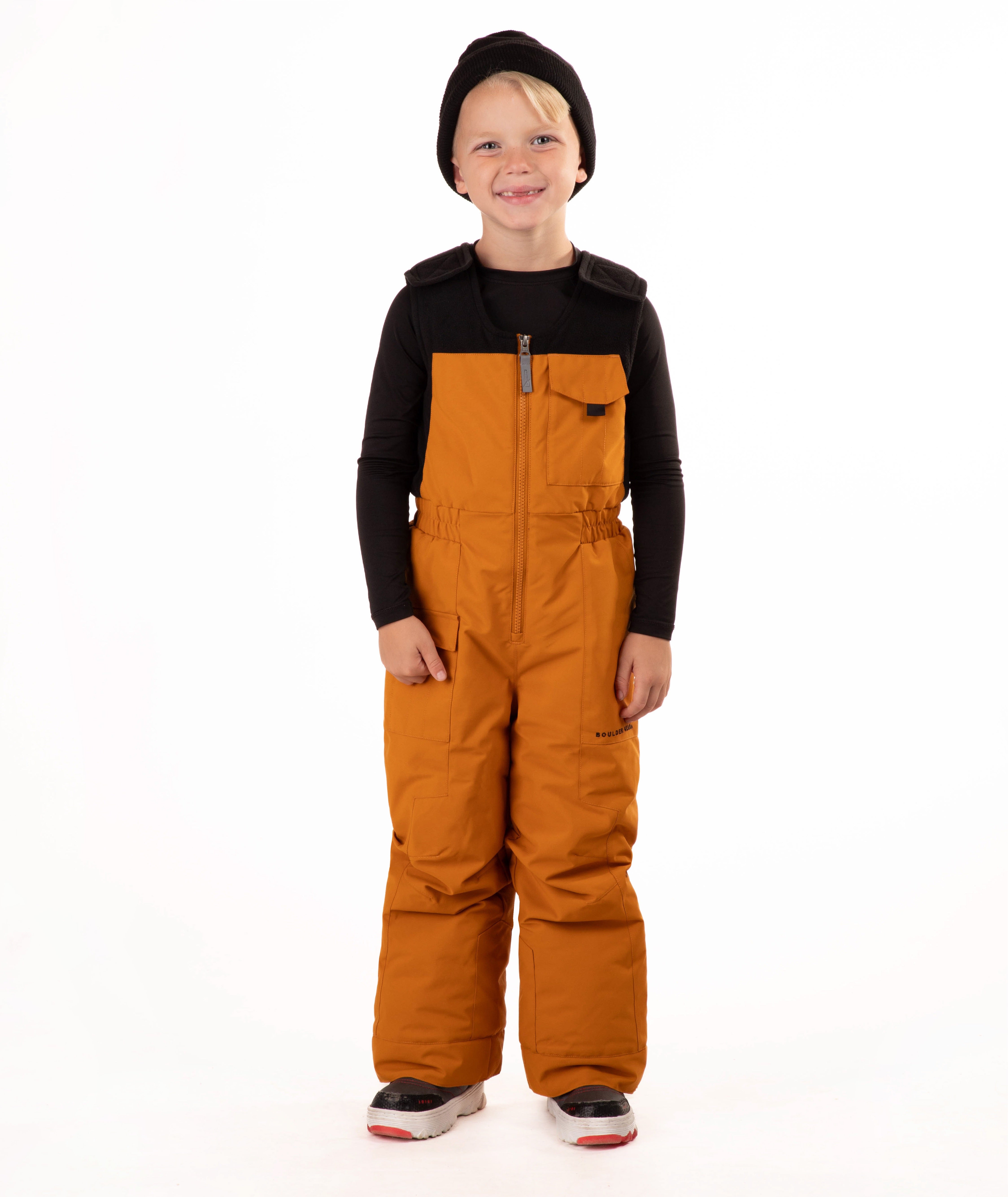 A child wearing a black knit beanie, a black long-sleeve top, and rust colored snow bib overalls, standing against a plain white background.