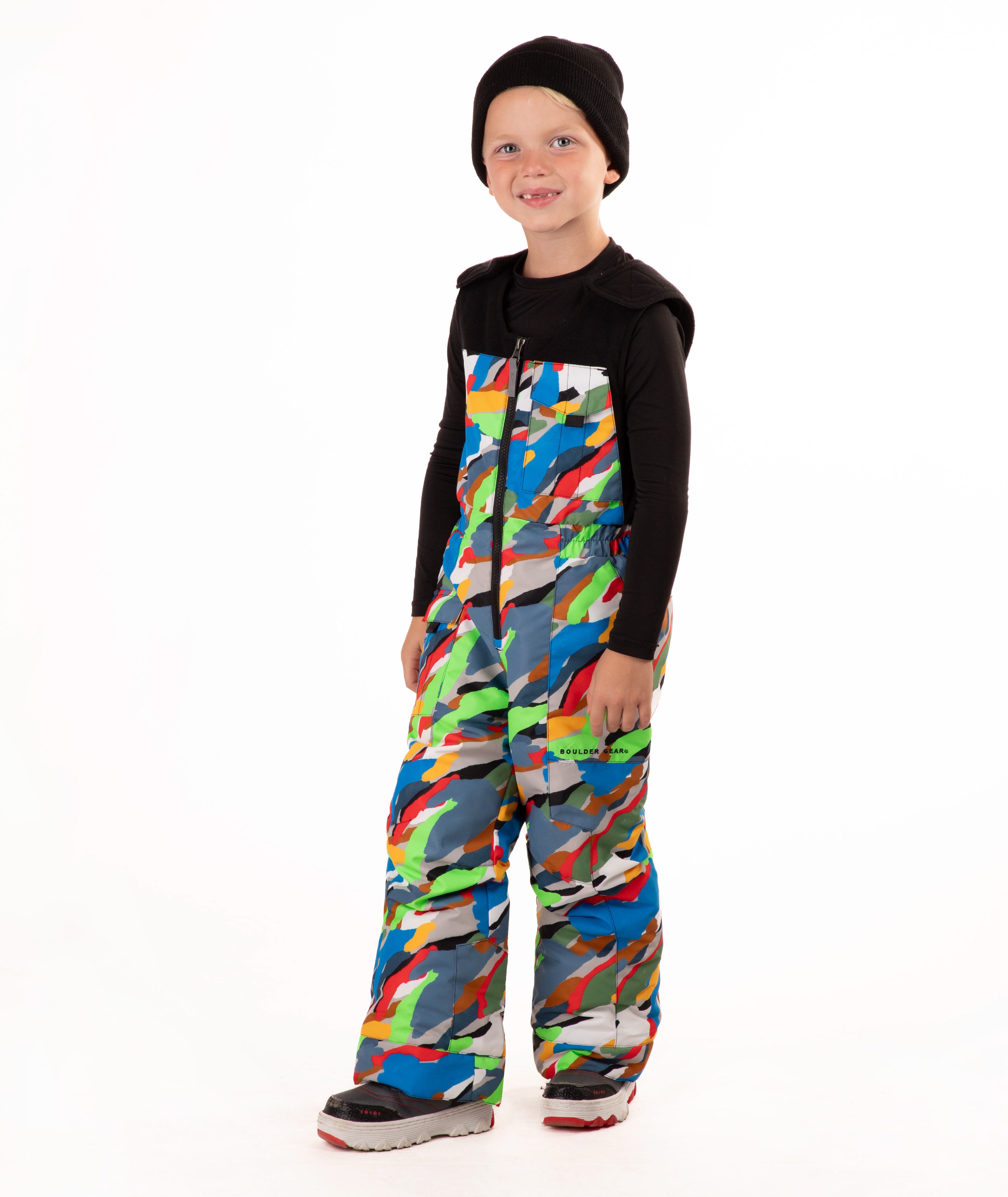 A child wearing a black knit beanie, a black long-sleeve top, and colorful camo print snow bib overalls, standing against a plain white background.