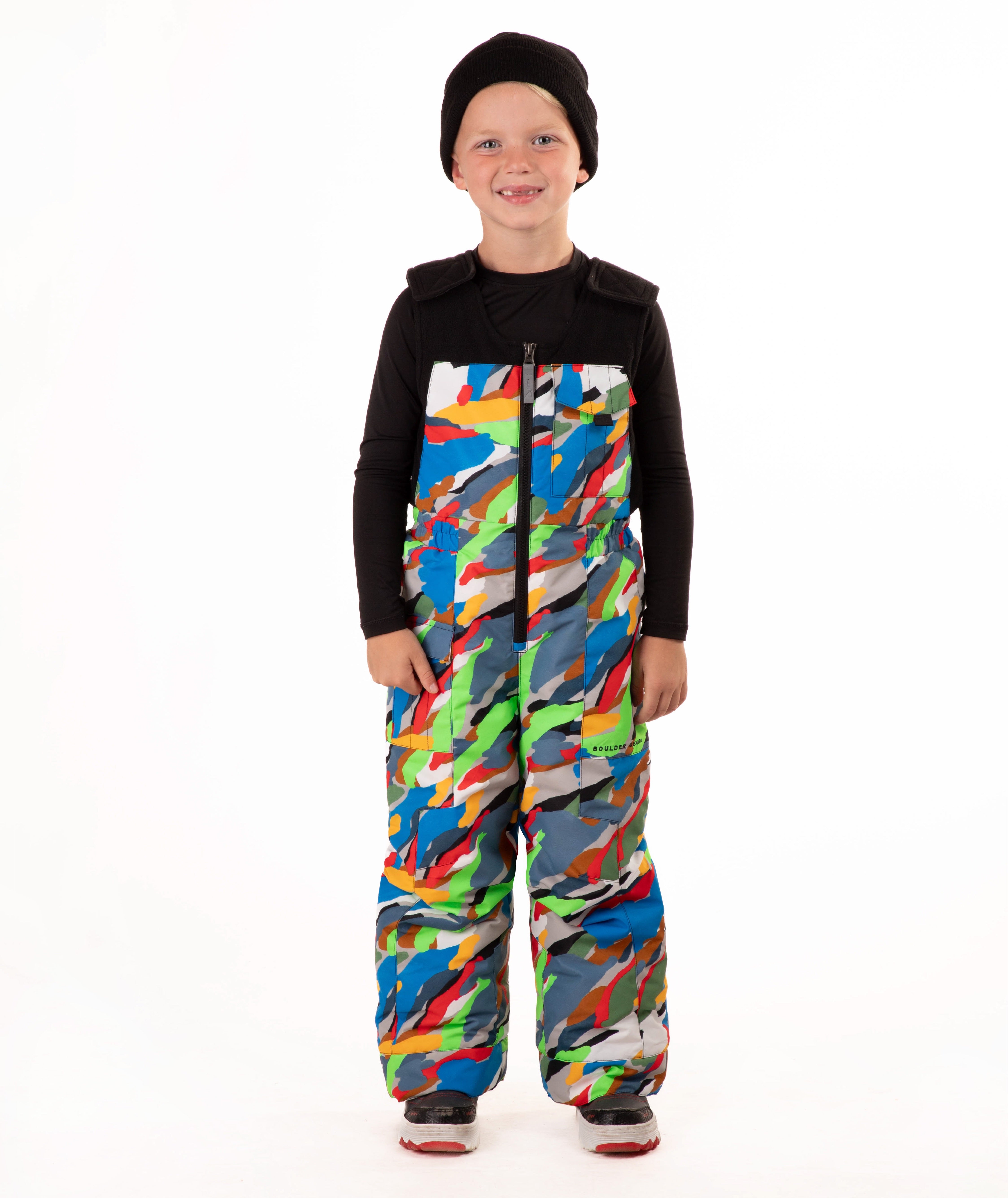 A child wearing a black knit beanie, a black long-sleeve top, and colorful camo print snow bib overalls, standing against a plain white background.