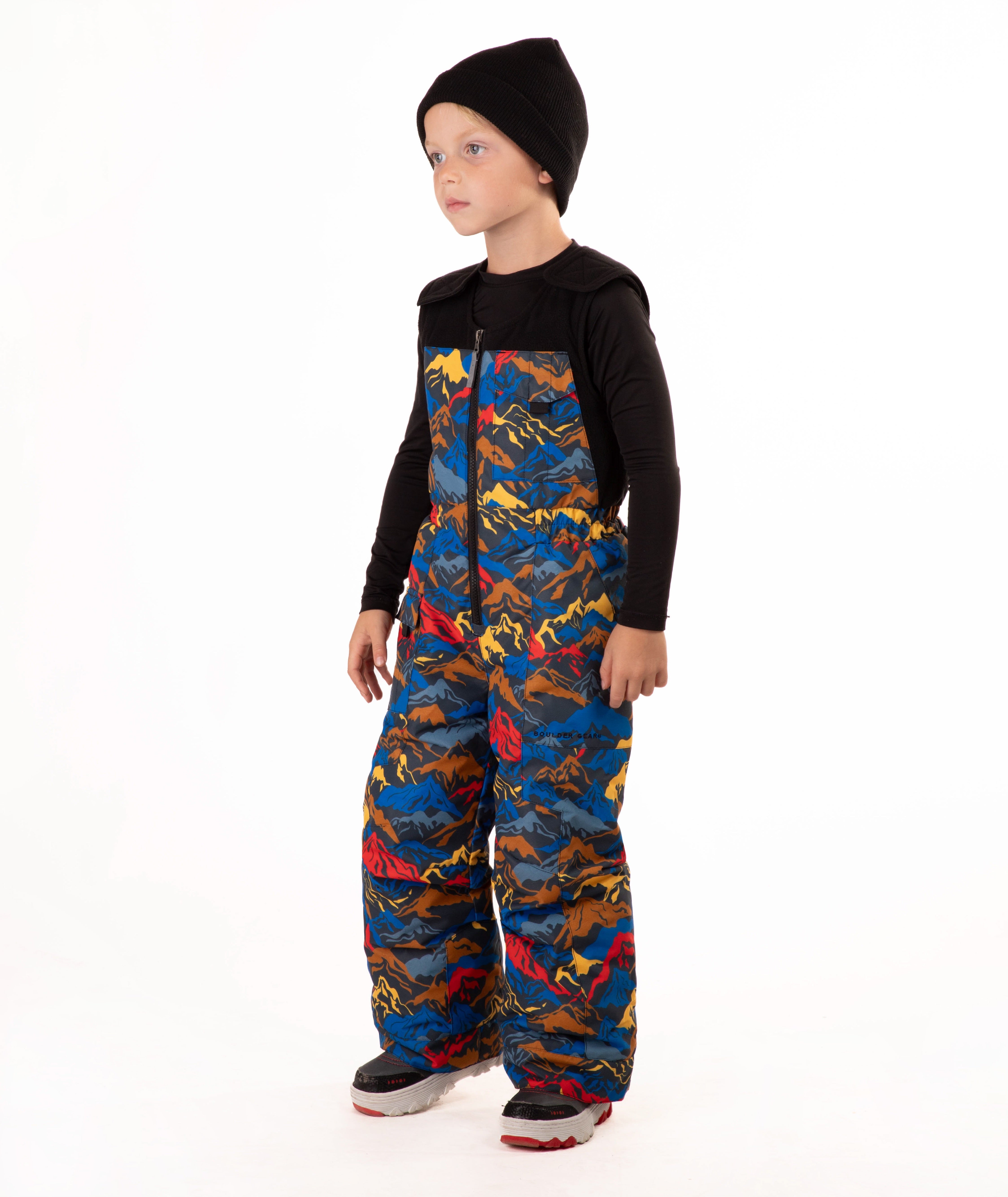 A child wearing a black knit beanie, a black long-sleeve top, and rust colored snow bib overalls, standing against a plain white background.