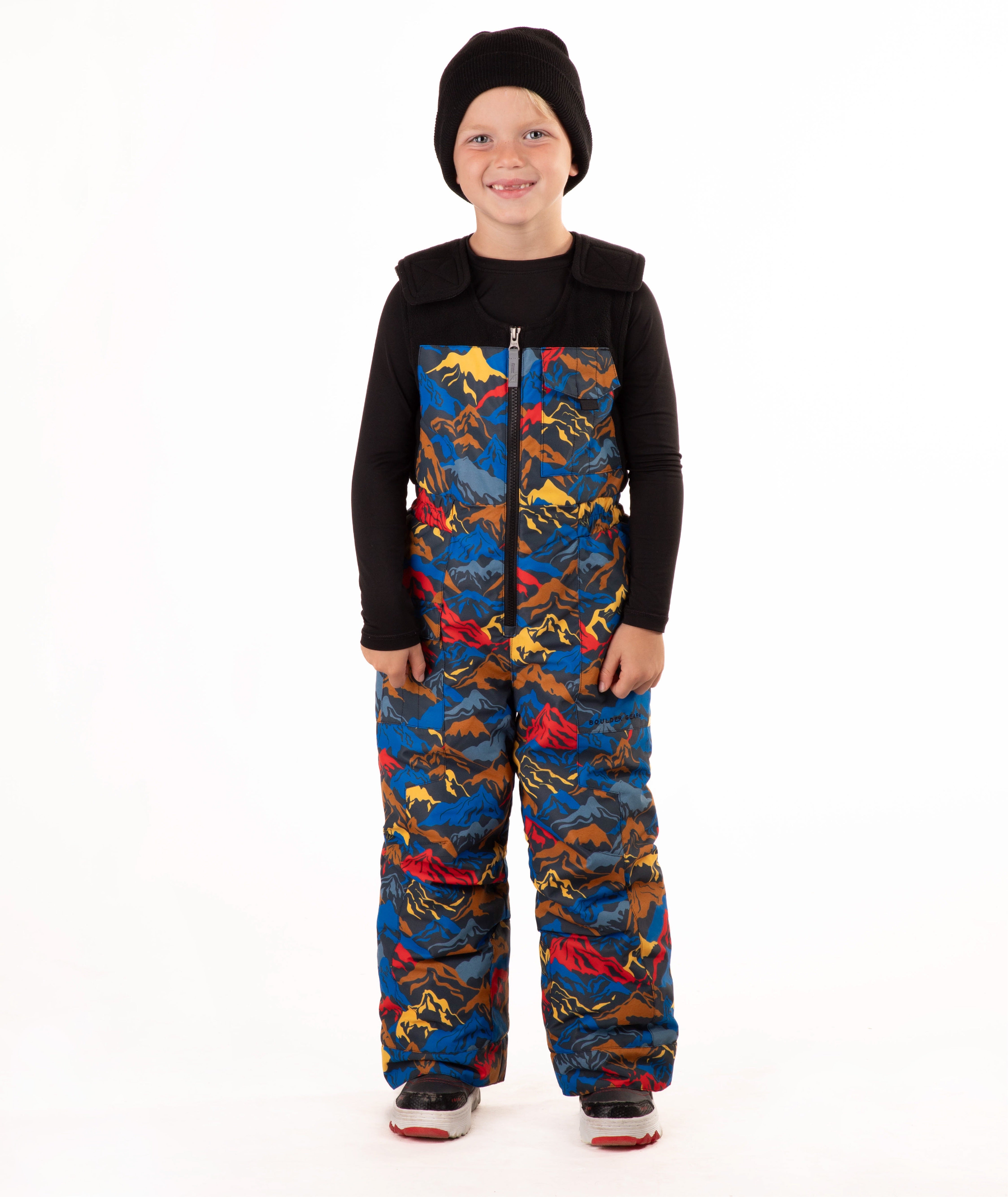 A child wearing a black knit beanie, a black long-sleeve top, and multi colored mountain print snow bib overalls, standing against a plain white background.