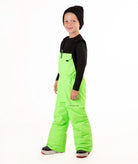 A child wearing bright neon green snow bib overalls over a black long-sleeve shirt and black beanie, standing against a plain white studio background.
