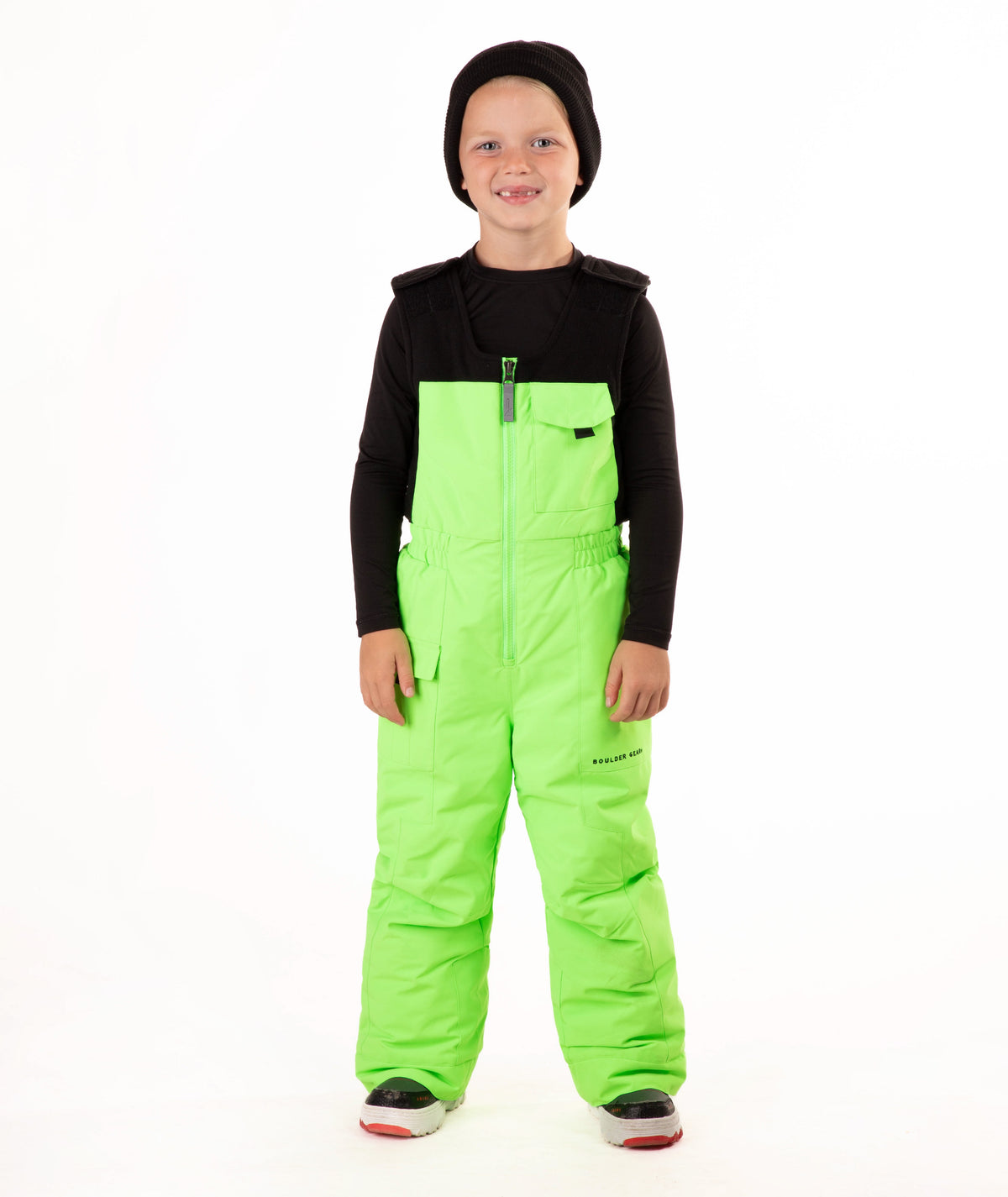 A child wearing bright neon green snow bib overalls over a black long-sleeve shirt and black beanie, standing against a plain white studio background.