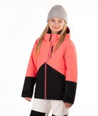 A young girl wearing a coral pink and black color-blocked ski jacket, a gray beanie, and off white ski pants, standing against a solid white background.