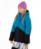 A young girl wearing a colorblock ski jacket in teal and black, a purple beanie, and colorful patterned ski pants, standing against a plain white background.