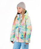 A young girl wearing a colorful tie-dye snowboard jacket, a gray beanie, and light blue snow pants, standing against a plain white background.