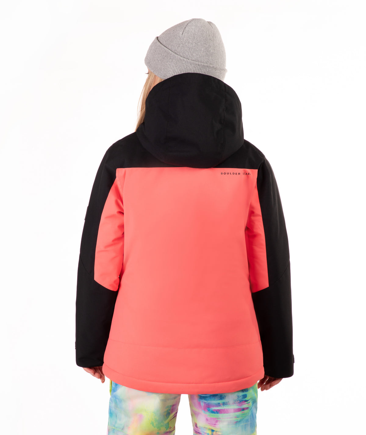 A person shown from the back wearing a color-blocked hooded jacket with a black hood and sleeves and a coral-pink body, paired with a gray beanie and tie-dye pants, against a plain white background.