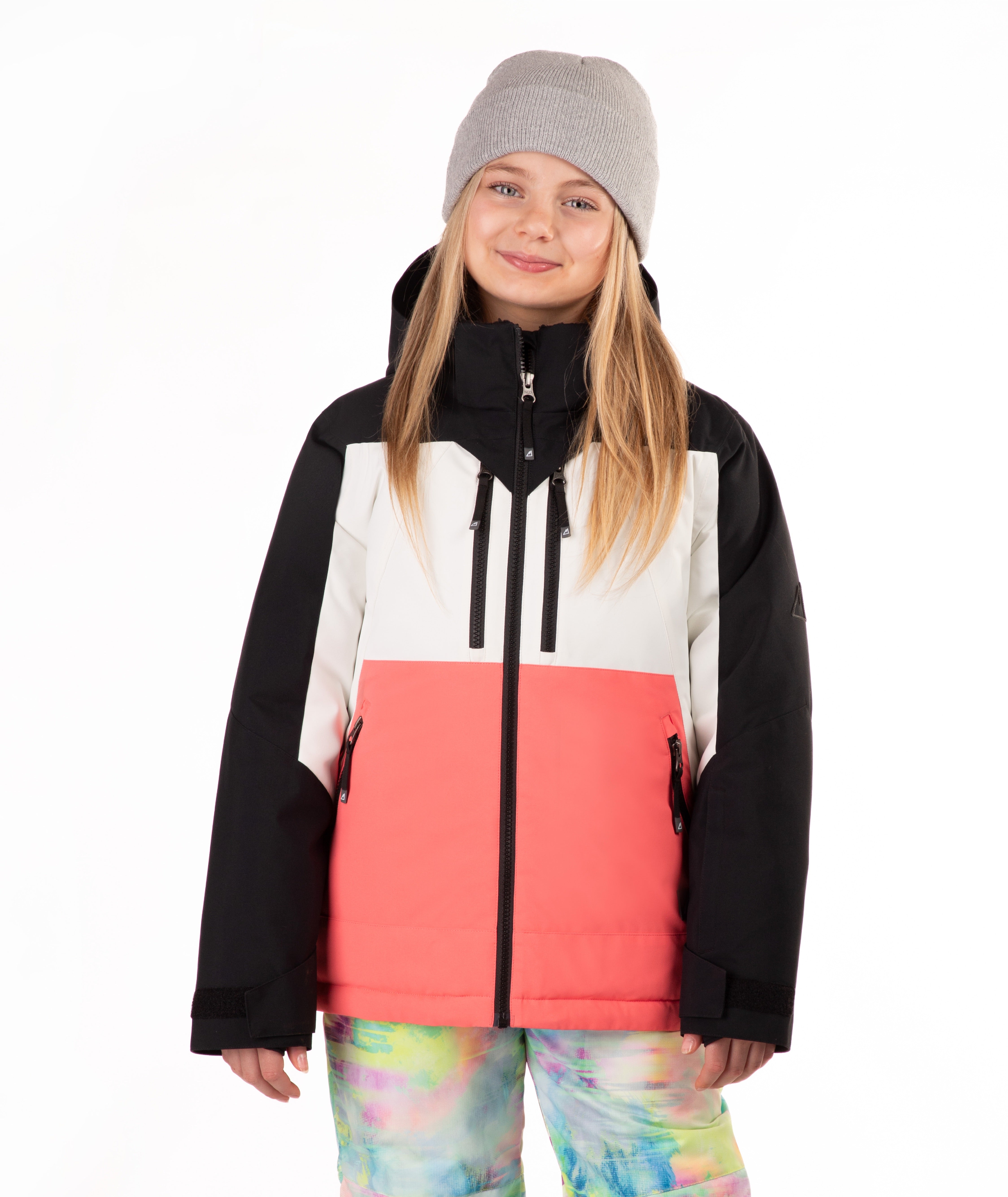 A young girl wearing a color-blocked ski jacket with black, white, and coral pink panels, a gray beanie, and tie-dye snow pants, photographed against a solid white background.