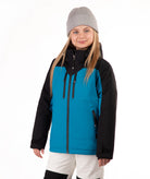 A young girl wearing a bright turquoise and black ski jacket, a gray beanie, and off white ski pants, standing against a plain white background.