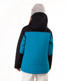 A young girl wearing a bright turquoise and black ski jacket, a gray beanie, and off white ski pants, standing against a plain white background.