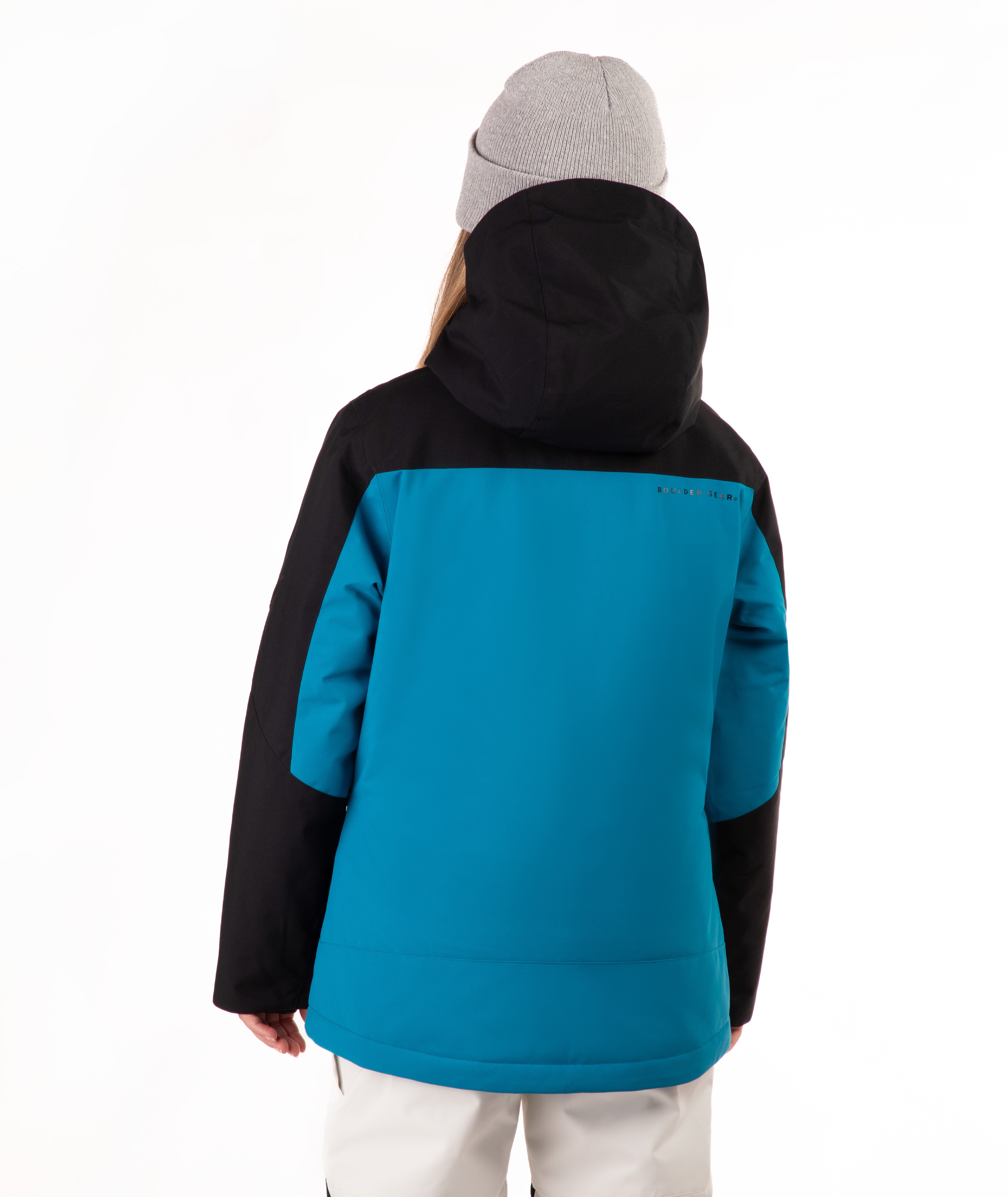 A young girl wearing a bright turquoise and black ski jacket, a gray beanie, and off white ski pants, standing against a plain white background.