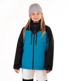 A young girl wearing a bright turquoise and black ski jacket, a gray beanie, and off white ski pants, standing against a plain white background.