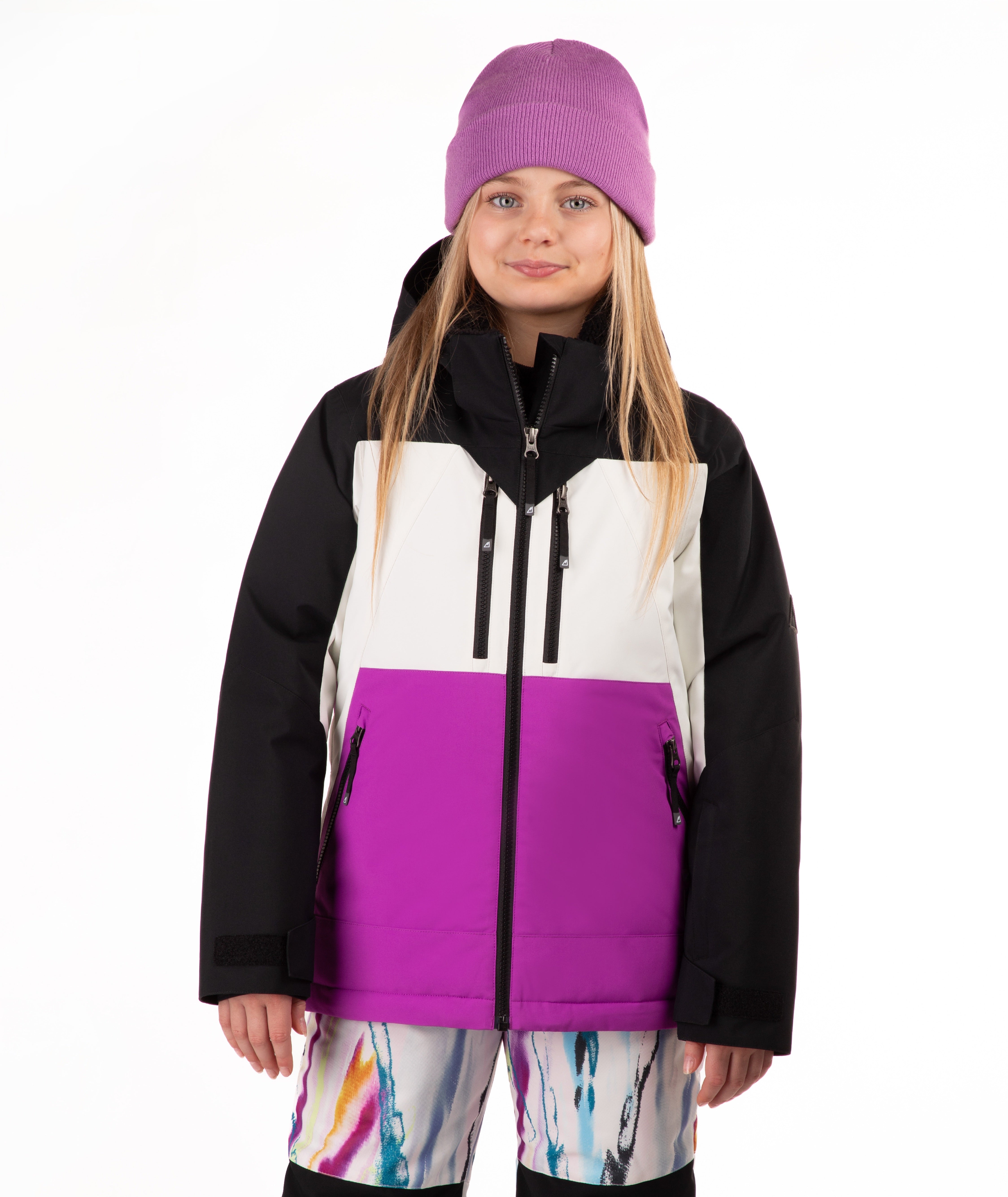 A young girl wearing a color-blocked ski jacket with black sleeves, a white upper chest, and a bright purple lower front panel, paired with a purple beanie and multicolor patterned snow pants, against a plain white background.