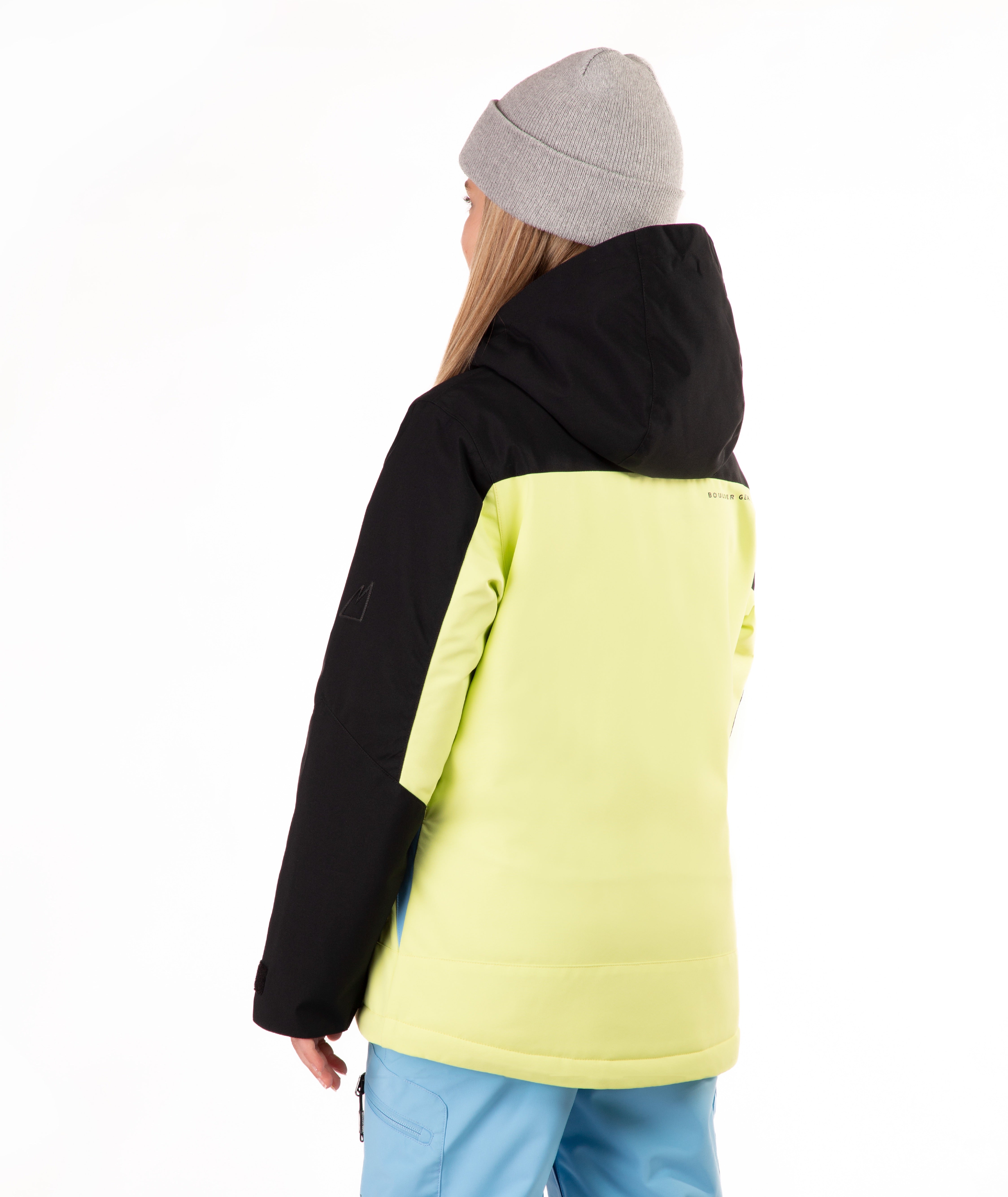 A model shown from the back wearing a color-blocked hooded jacket with a black upper section and a bright neon yellow lower section, paired with a gray beanie and light blue pants, against a plain white background.