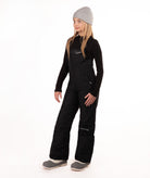 A young girl modeling black snow bib overalls with a gray beanie, black long-sleeve shirt, and gray boots against a white studio background.