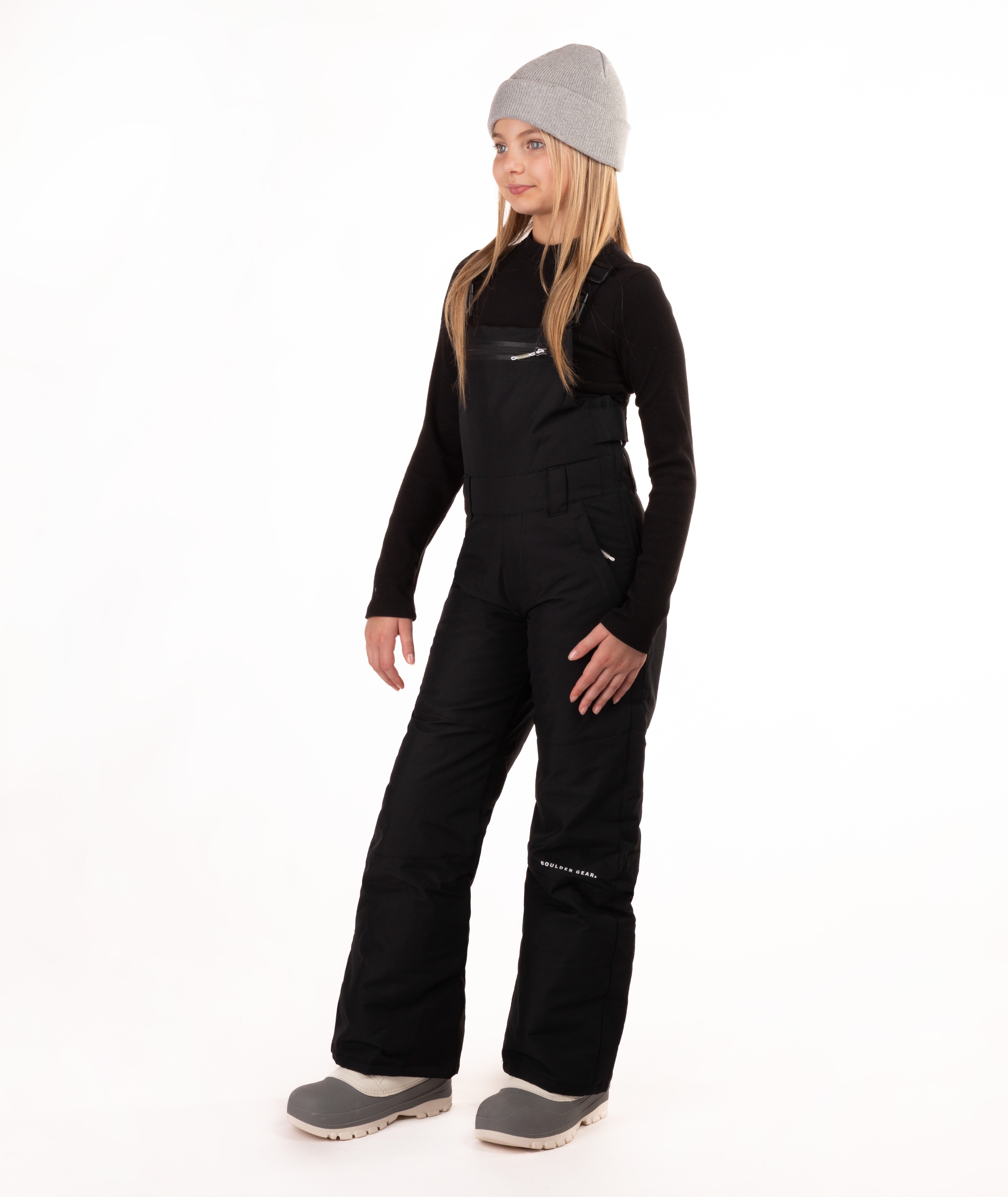 A young girl modeling black snow bib overalls with a gray beanie, black long-sleeve shirt, and gray boots against a white studio background.